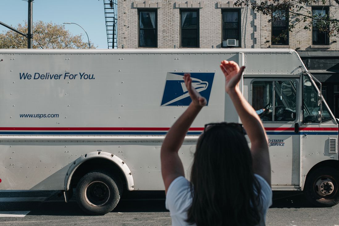 A woman cheers on a passing USPS truck, which says "We deliver for you"